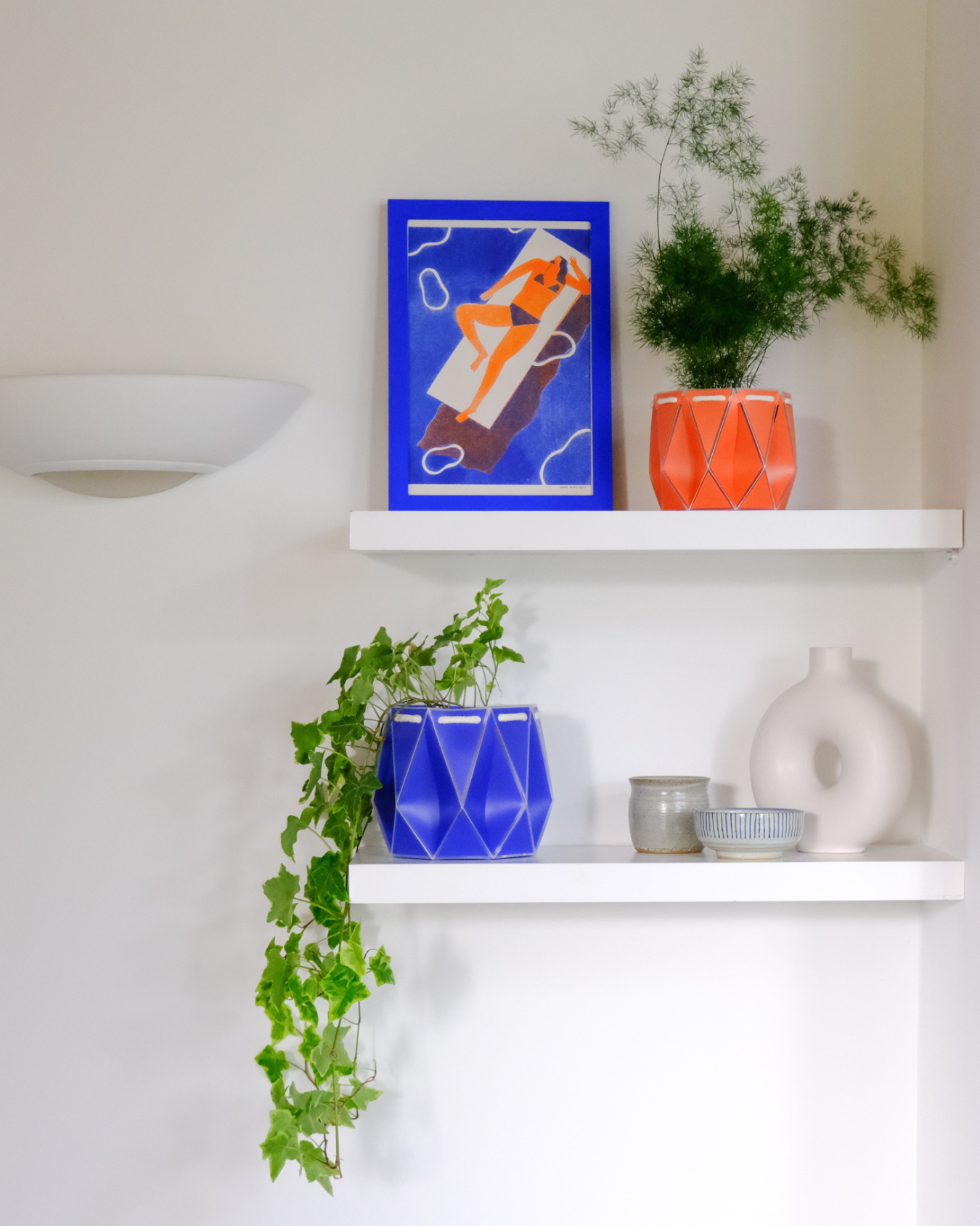 Two minimalist white shelves mounted on a white wall, holding colorful origami-style POTR self-watering plant pots. The top shelf displays a coral POTR pot with an airy green plant and a framed illustration of a sunbather. The bottom shelf features a blue POTR pot with trailing ivy, accompanied by ceramics and a white sculptural vase.